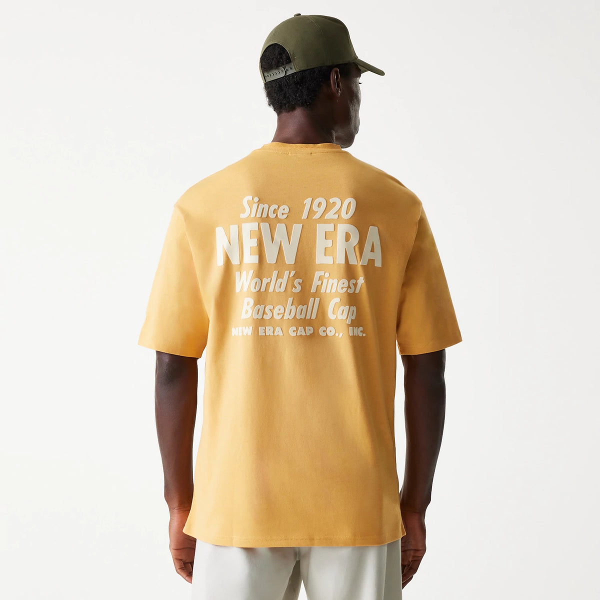 New Era Graphic Beige Oversized T-Shirt, rear view showing a large white printed graphic with Since 1920 New Era World's Finest Baseball Cap text on the upper back, worn by a person with a green cap.