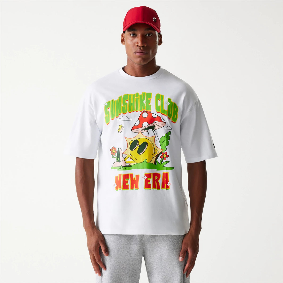 New Era Outdoor Graphic White Oversized T-Shirt, front view showing a colourful graphic with a sun in sunglasses under a mushroom, with Sunshine Club above and New Era below in bright letters, worn by a person in a red cap and grey trousers.