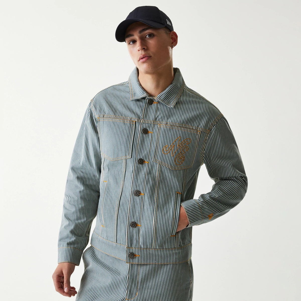 New Era Blue Denim Jacket, front view showing vertical stripes with button closures down the centre, an orange raised logo on the left chest, and contrasting stitching, worn by a model with a dark cap.