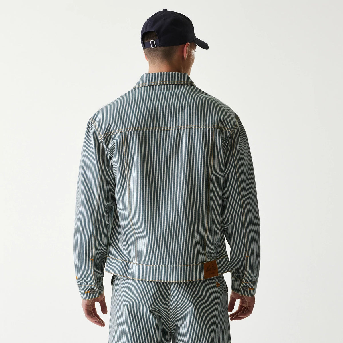 New Era Blue Denim Jacket, rear view showing the blue denim striped pattern with a pointed collar, straight shoulder seam, and a brown leather patch sewn at the lower right hem, worn by a model in matching trousers and a navy cap.