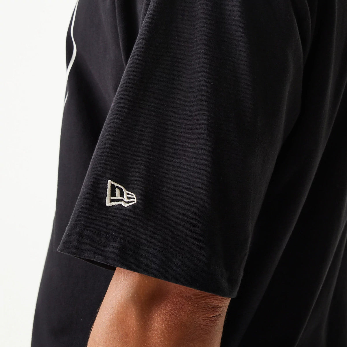 New Era Crest Black Oversized T-Shirt, side view showing the black short sleeve with a raised New Era logo embroidered near the hem, highlighting the relaxed fit and length of the sleeve.