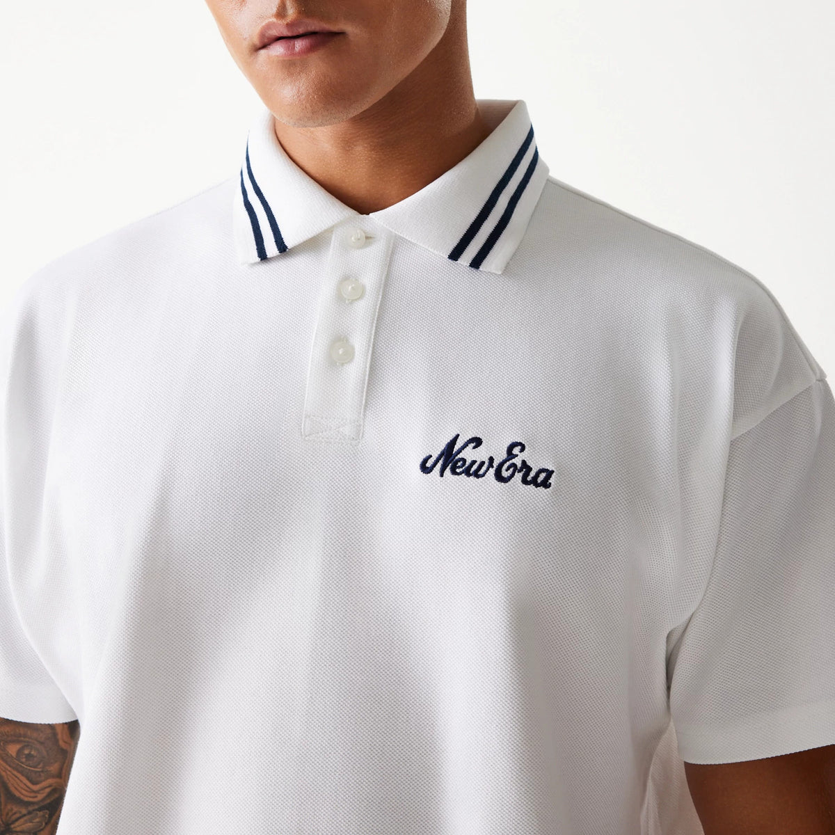 New Era Drop Shoulder White Polo Shirt, front view on a model showing the embroidered New Era logo in navy on the left chest, with a three-button placket and navy double stripe detail on the collar.