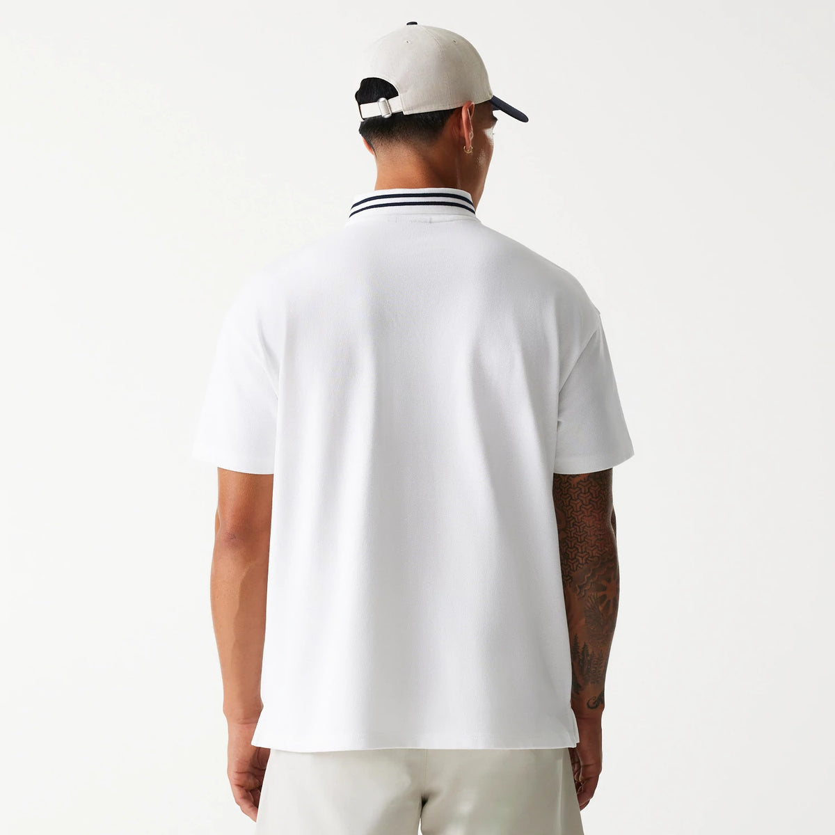 New Era Drop Shoulder White Polo Shirt, back view on a model wearing a beige cap, displaying the shirt's relaxed fit and contrast stripe detail on the collar, coupled with light-coloured shorts.