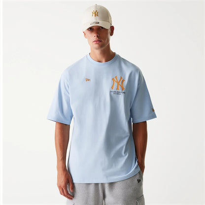 New Era New York Yankees MLB Baseball Graphic Pastel Blue Oversized T-Shirt, front view featuring a large gold New York Yankees logo on the chest, a small debossed New Era logo on the upper left, and a matching cap worn by the model.