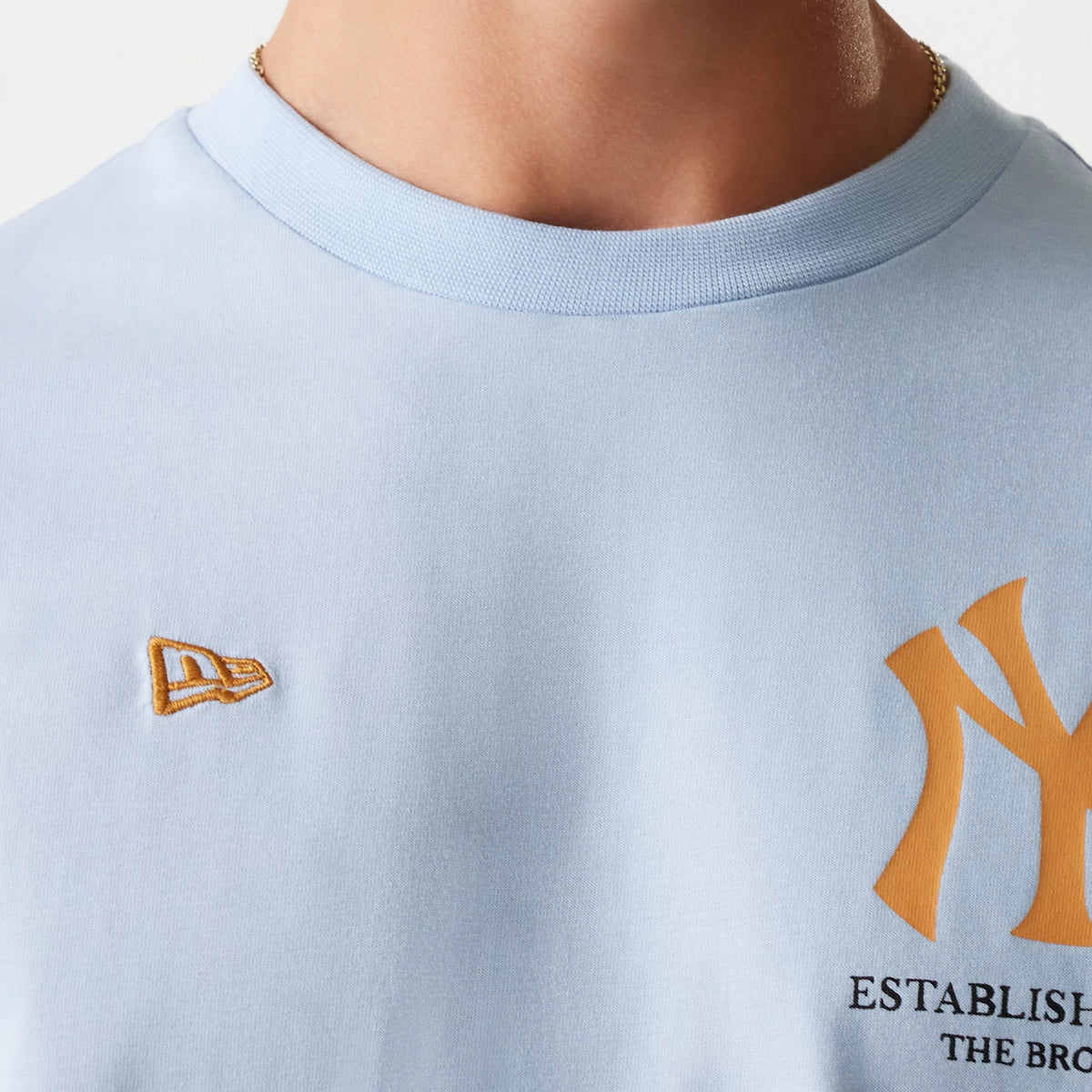 New Era New York Yankees MLB Baseball Graphic Pastel Blue Oversized T-Shirt, front view showing a light blue fabric with a raised yellow New Era logo embroidered on the left chest and a large yellow Yankees logo on the right side.