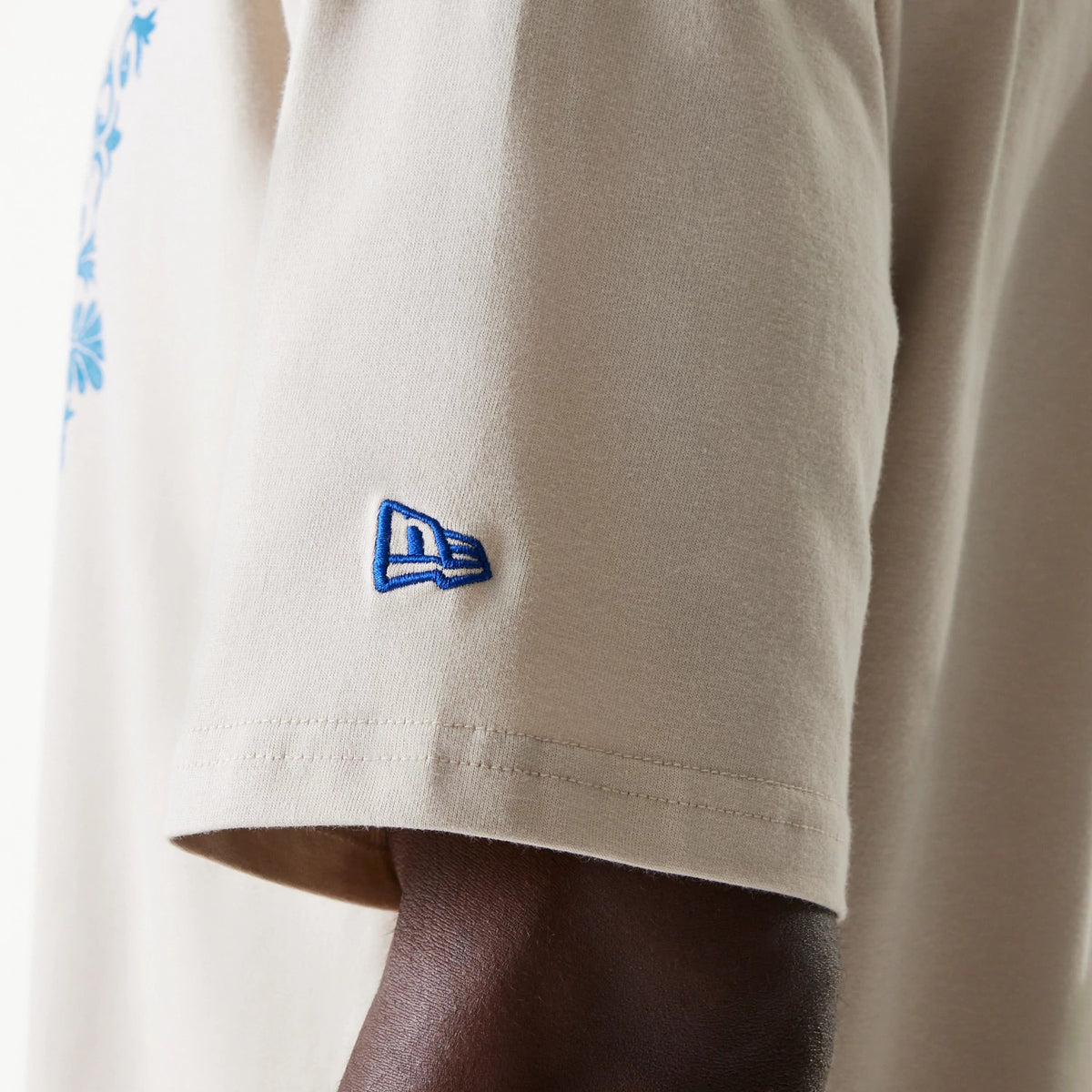 New Era LA Dodgers MLB Floral Graphic Cream Oversized T-Shirt, side view showing a blue embroidered New Era logo on the left sleeve, part of a light cream short sleeve with a glimpse of blue floral pattern on the main body.