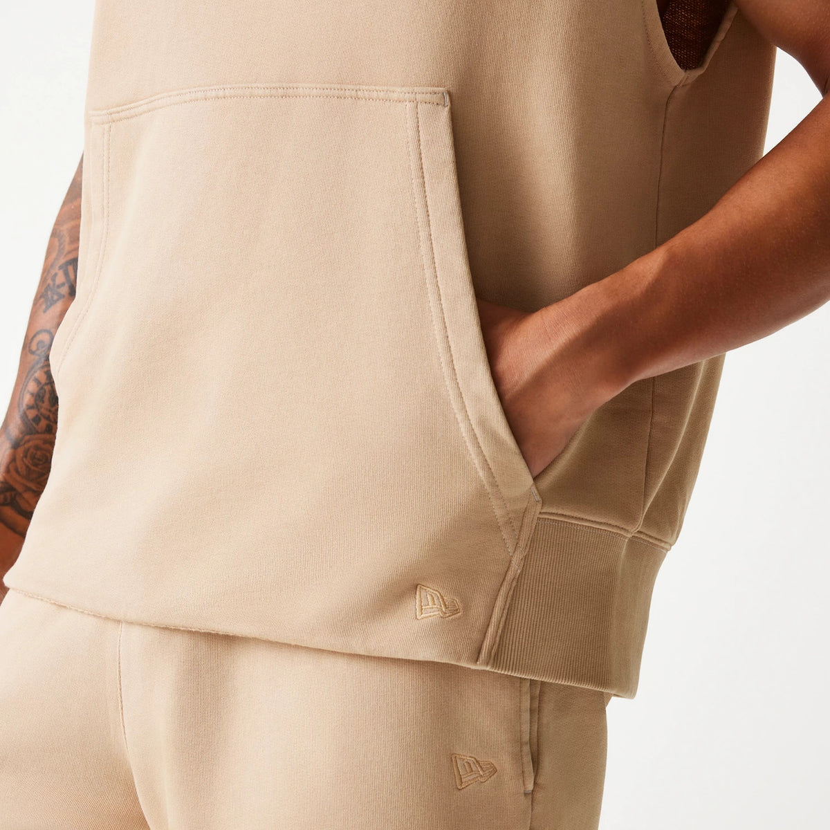 New Era Chicago Bulls NBA Washed Beige Sleeveless Hoodie, side view showing a hand in the large front pocket of the beige hoodie, with a debossed New Era logo near the ribbed hem and matching beige shorts.