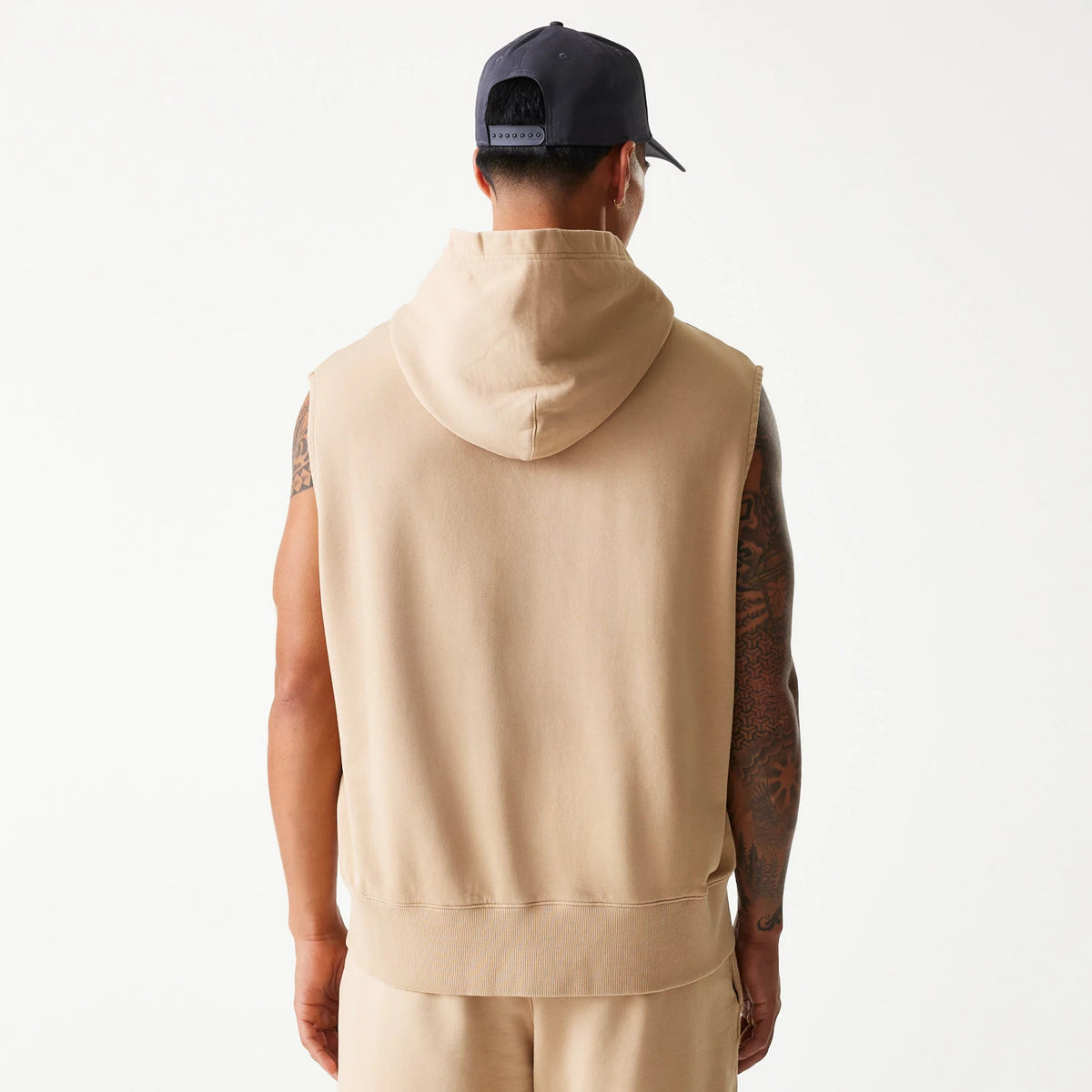 New Era Chicago Bulls NBA Washed Beige Sleeveless Hoodie, rear view showing the plain beige back with a relaxed hood, sleeveless cut, and straight hem, worn by an individual also wearing a dark cap with eyelets and an adjustable strap at the back.