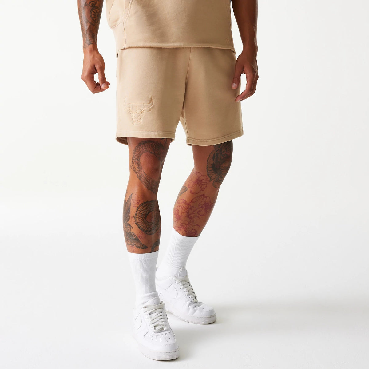 New Era Chicago Bulls NBA Washed Beige Shorts, on-model view showing the above-knee length shorts in a washed beige colour with an embossed Chicago Bulls logo on the left thigh, paired with white socks and sneakers.