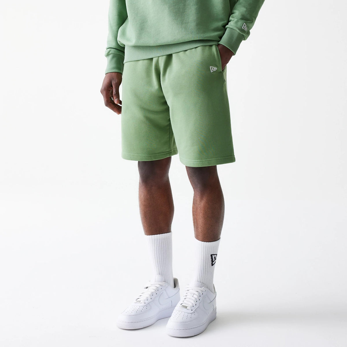 New Era Essential Green Oversized Shorts, on-model view showing the relaxed green shorts with a raised New Era logo on the left thigh, paired with matching green top and white trainers, highlighting a casual outfit style.