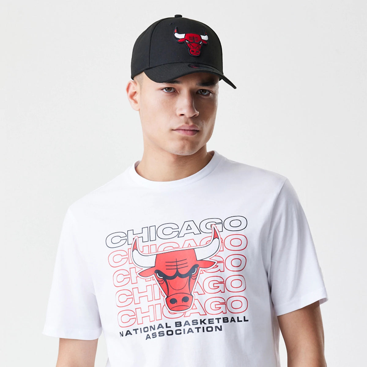 New Era Chicago Bulls NBA Stack Wordmark White T-Shirt, on-model view featuring a large red Bulls logo centred amidst repeated Chicago text, worn with a black cap showing an embossed Bulls logo on the front panel.