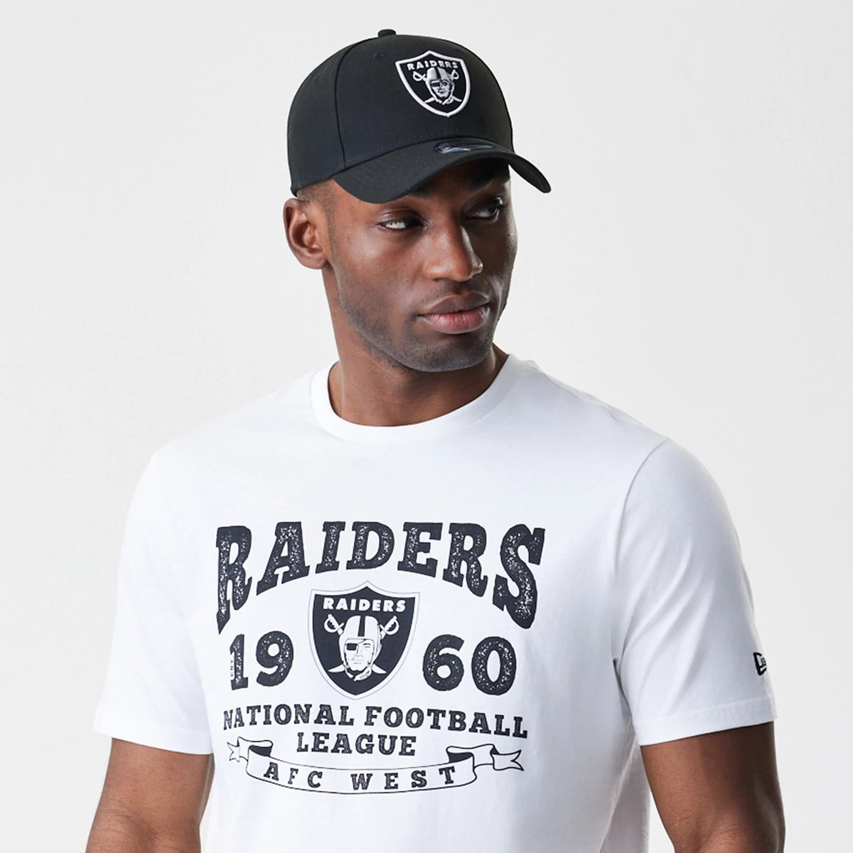 New Era Las Vegas Raiders NFL Stack Arch Logo White T-Shirt, on-model view showing black printed Raiders text, the football helmet logo centred on the chest, and a debossed New Era logo on the side of a black cap worn by the model.