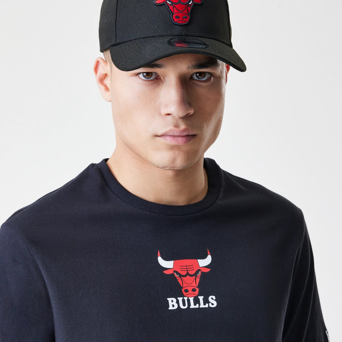 New Era Chicago Bulls NBA Logo Black T-Shirt, on-model view showing a black T-shirt featuring a printed red Chicago Bulls logo and the word BULLS on the chest, paired with a black cap with a raised Bulls logo on the front.