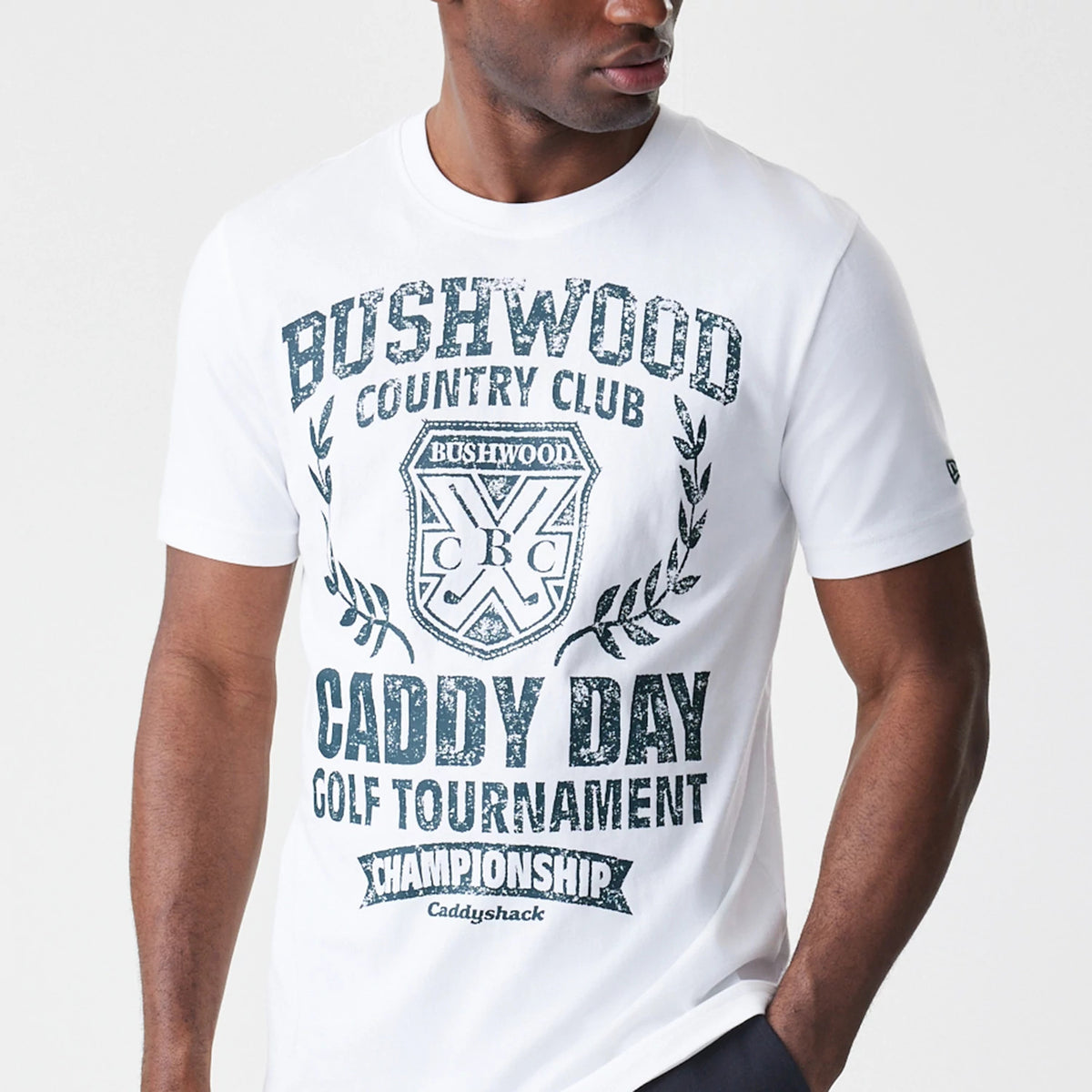 New Era Caddyshack Caddy Day Tournament White T-Shirt, on-model view showing a large distressed blue graphic on the front with 'Bushwood Country Club' and 'Caddy Day Golf Tournament', a small New Era logo on the left sleeve.