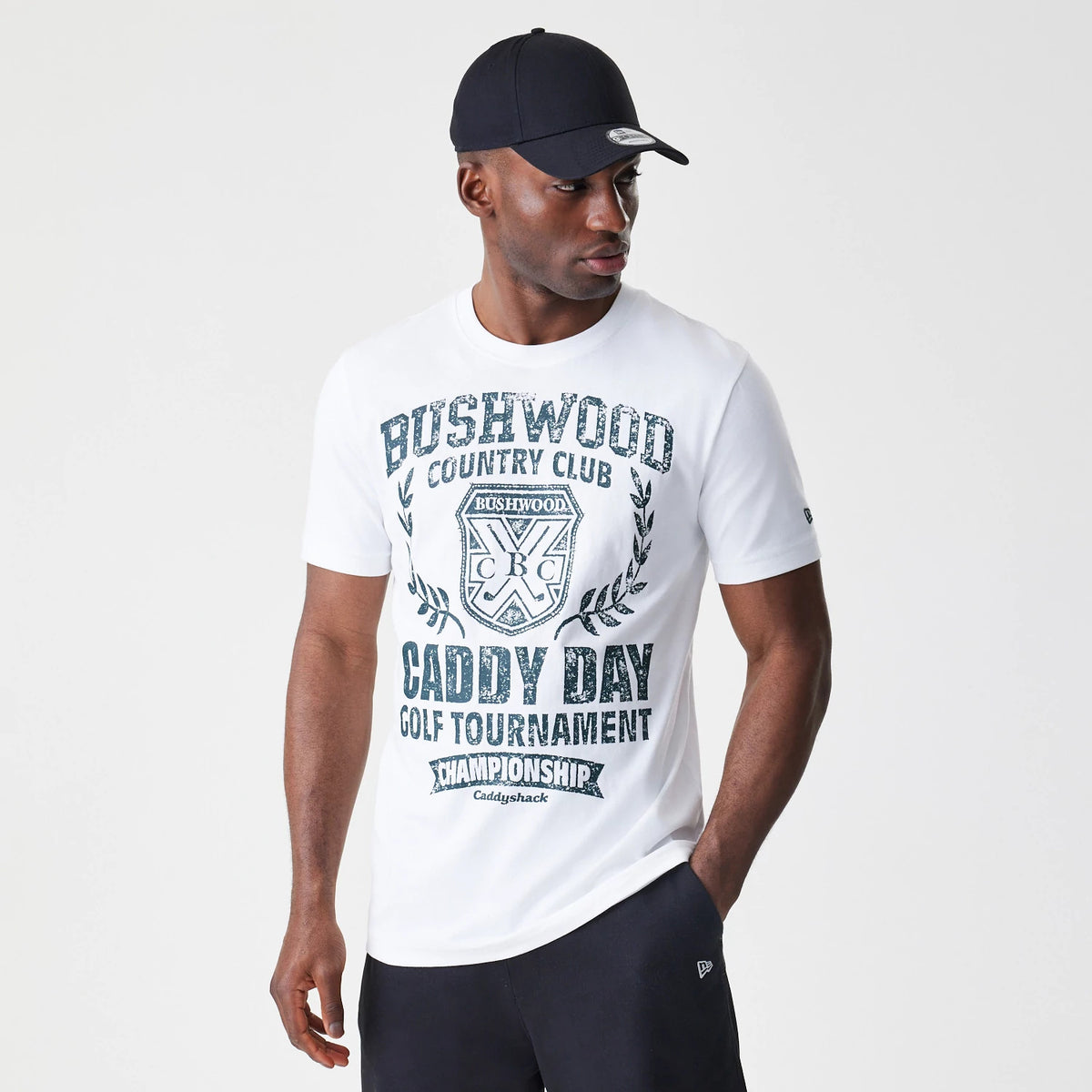 New Era Caddyshack Caddy Day Tournament White T-Shirt, on-model view showing a man wearing the white T-shirt with a bold graphic reading Bushwood Country Club Caddy Day Golf Tournament and a New Era logo on the sleeve, paired with black trousers.