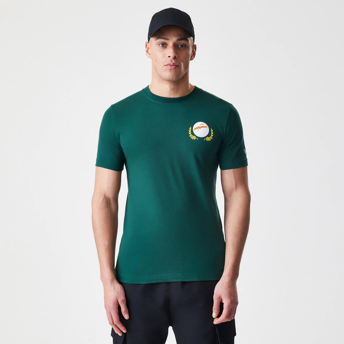 New Era Caddyshack It's In The Hole Dark Green T-Shirt, on-model view showing the dark green shirt with a round logo on the left chest and New Era logo on the left sleeve, paired with a black cap on the model.