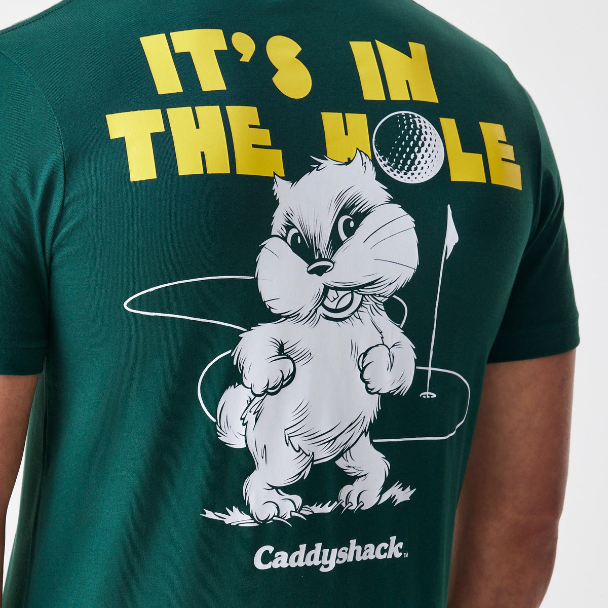 New Era Caddyshack It's In The Hole Dark Green T-Shirt, rear view showing large yellow text at the top, a white cartoon gopher holding a golf flag beneath, and the word Caddyshack printed in white at the bottom.