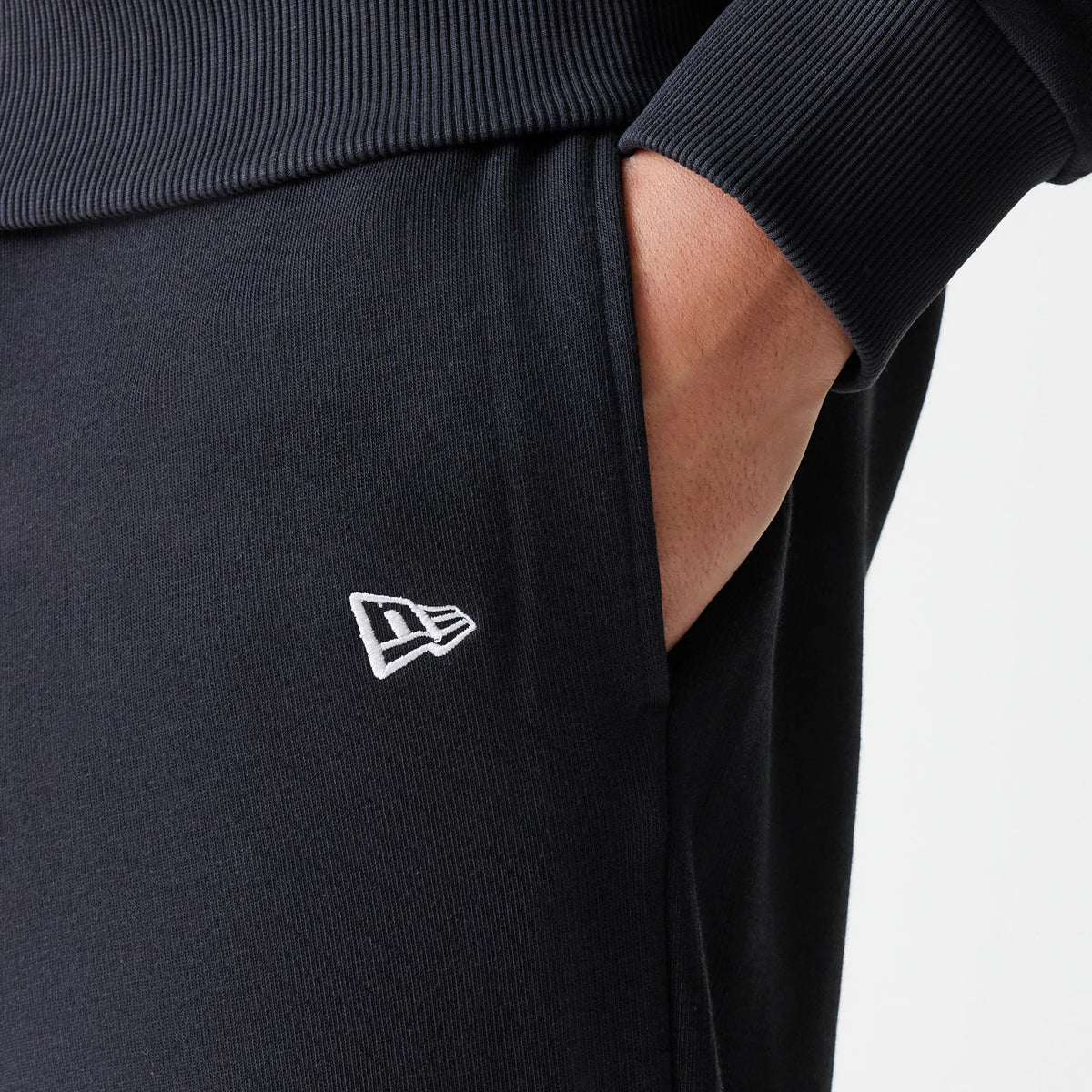 New Era New York Yankees MLB League Essential Midi French Terry Black Joggers, close-up showing the white embroidered New Era flag logo near the side pocket, with ribbed waistband and hand partially inside the pocket.
