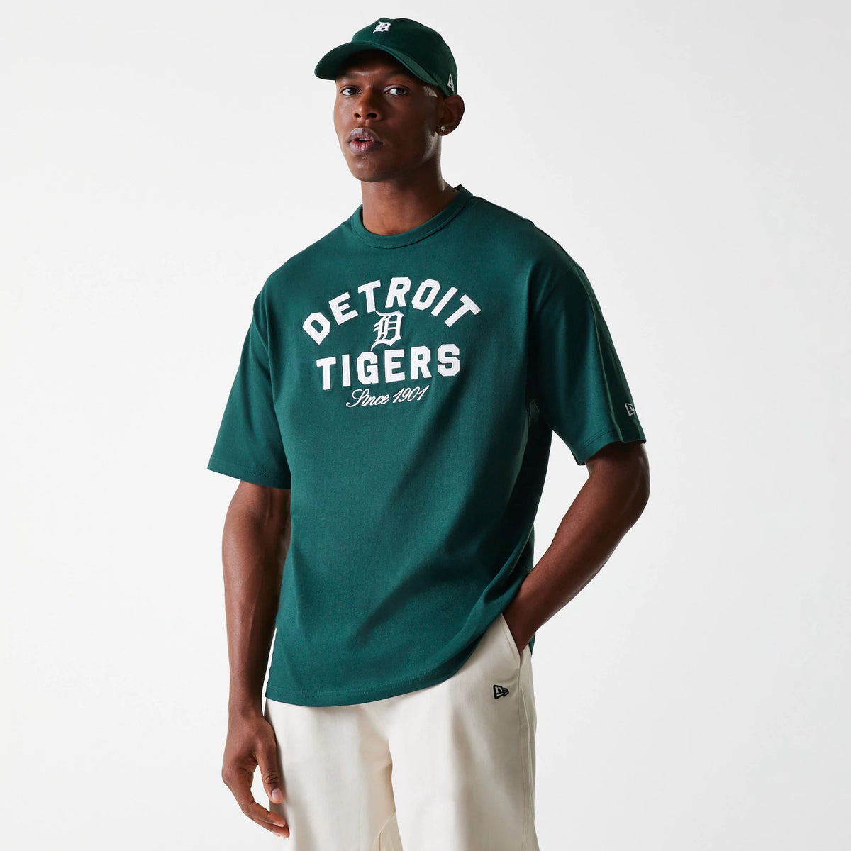 New Era Detroit Tigers MLB Heritage Dark Green Oversized T-Shirt, on-model view showing a dark green T-shirt with white printed team graphics on the chest, worn by a model in matching cap and light trousers with a debossed logo on the left sleeve.