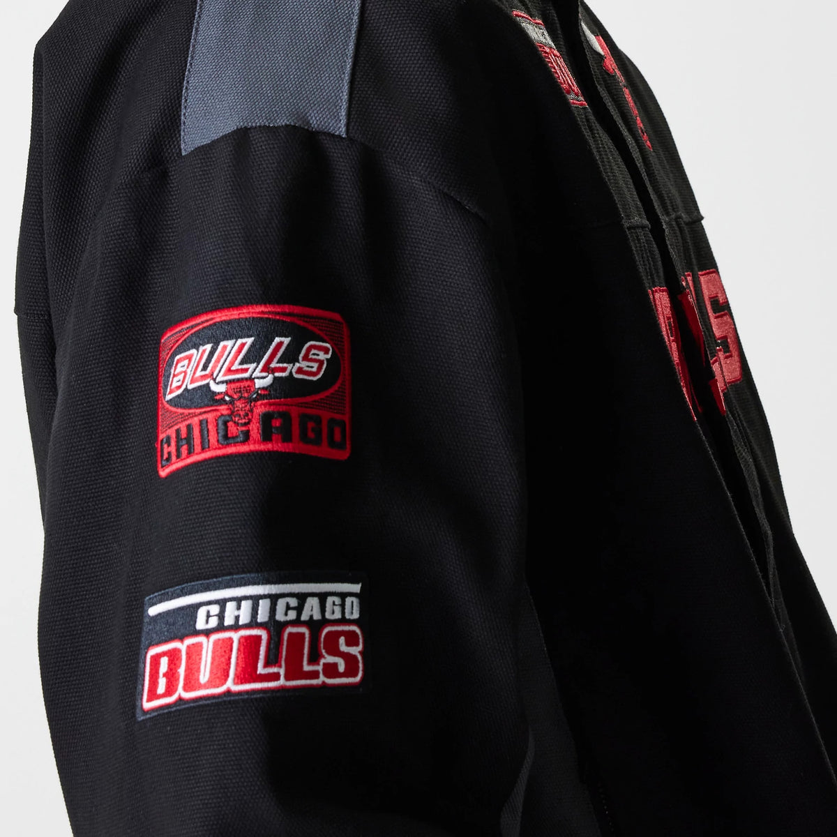 New Era Chicago Bulls NBA Black Motorsports Jacket, side view showing red and white embroidered Bulls patches on the sleeve, a grey panel on the shoulder, and part of the red Bulls logo on the black body of the jacket.
