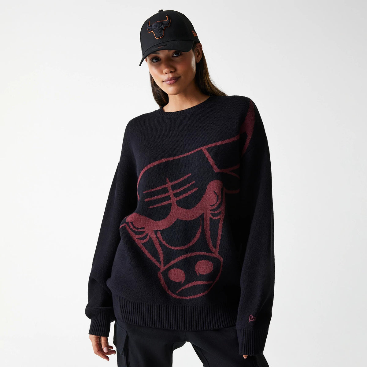 New Era Chicago Bulls NBA Knit Black Oversized Knit Sweater, on a model with a matching black cap, features a large red Chicago Bulls logo knitted across the front, complemented by a New Era flag logo on the left sleeve.