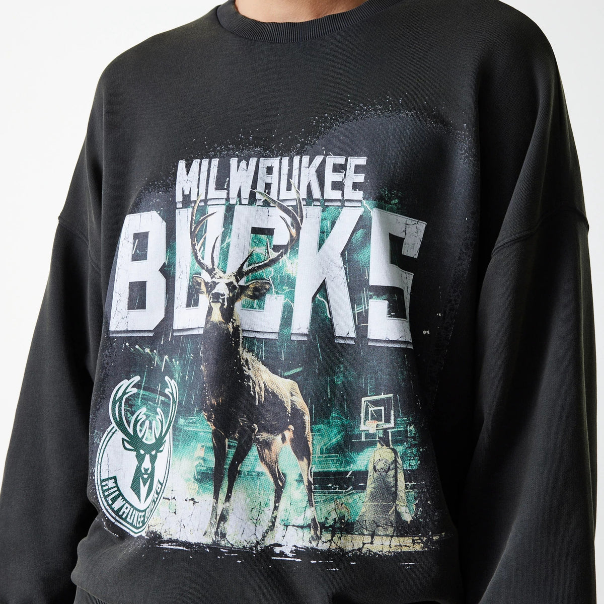 New Era Milwaukee Bucks NBA Washed Graphic Grey Oversized Crew Neck Sweater, front view on a model featuring a large distressed Bucks logo and a detailed deer graphic with a basketball hoop in the background.