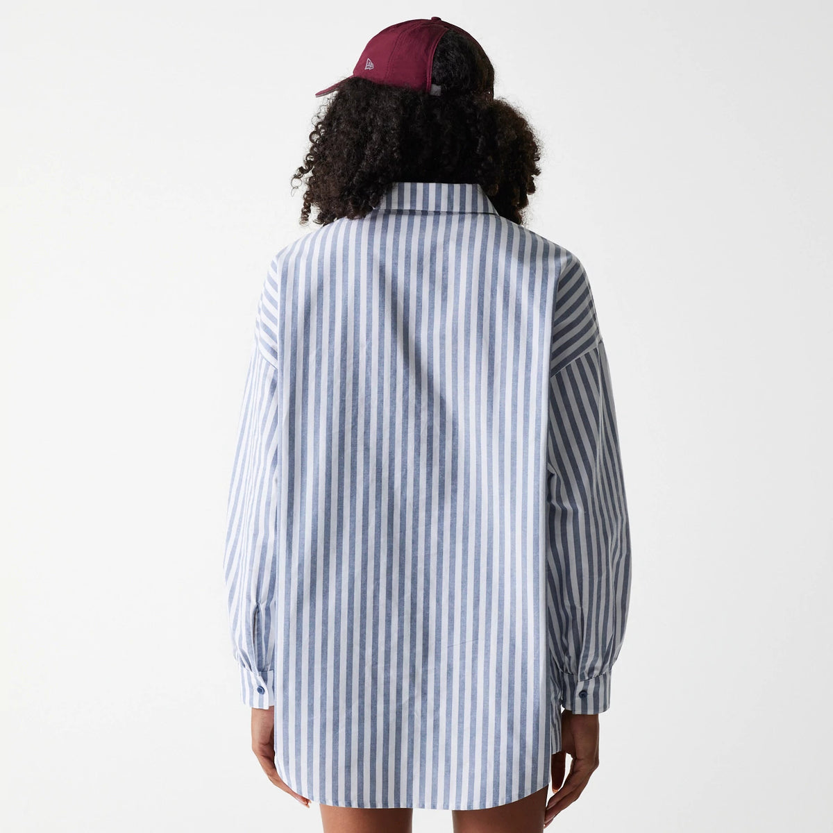 New Era New York Yankees Womens MLB Stripe Dark Blue Long Sleeve Shirt, back view on a model with a curly hairstyle and a burgundy cap, showcasing vertical blue and white stripes and a rounded hem.