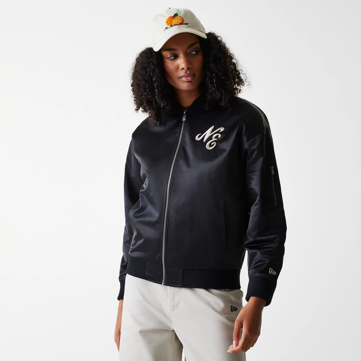 Womens New Era Black Satin Bomber Jacket, on-model view showing the black zip-up jacket with a large embroidered NE logo on the left chest, side pocket zip, ribbed cuffs and hem, and a small New Era logo on the sleeve.
