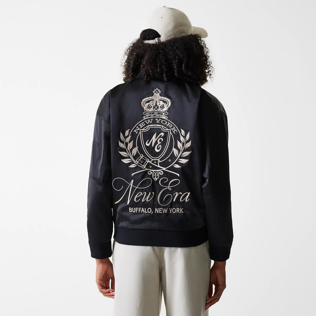 Womens New Era Black Satin Bomber Jacket, rear view showing a large embossed emblem with a crown and laurel design, and New Era Buffalo, New York text, worn by a person in light trousers and a cream cap.
