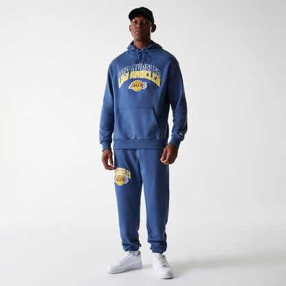 New Era LA Lakers NBA Arch Graphic Dark Blue Joggers, on a model with a matching hoodie, showing the LA Lakers logo embroidered in yellow and white on the left thigh, paired with white trainers and a black cap.