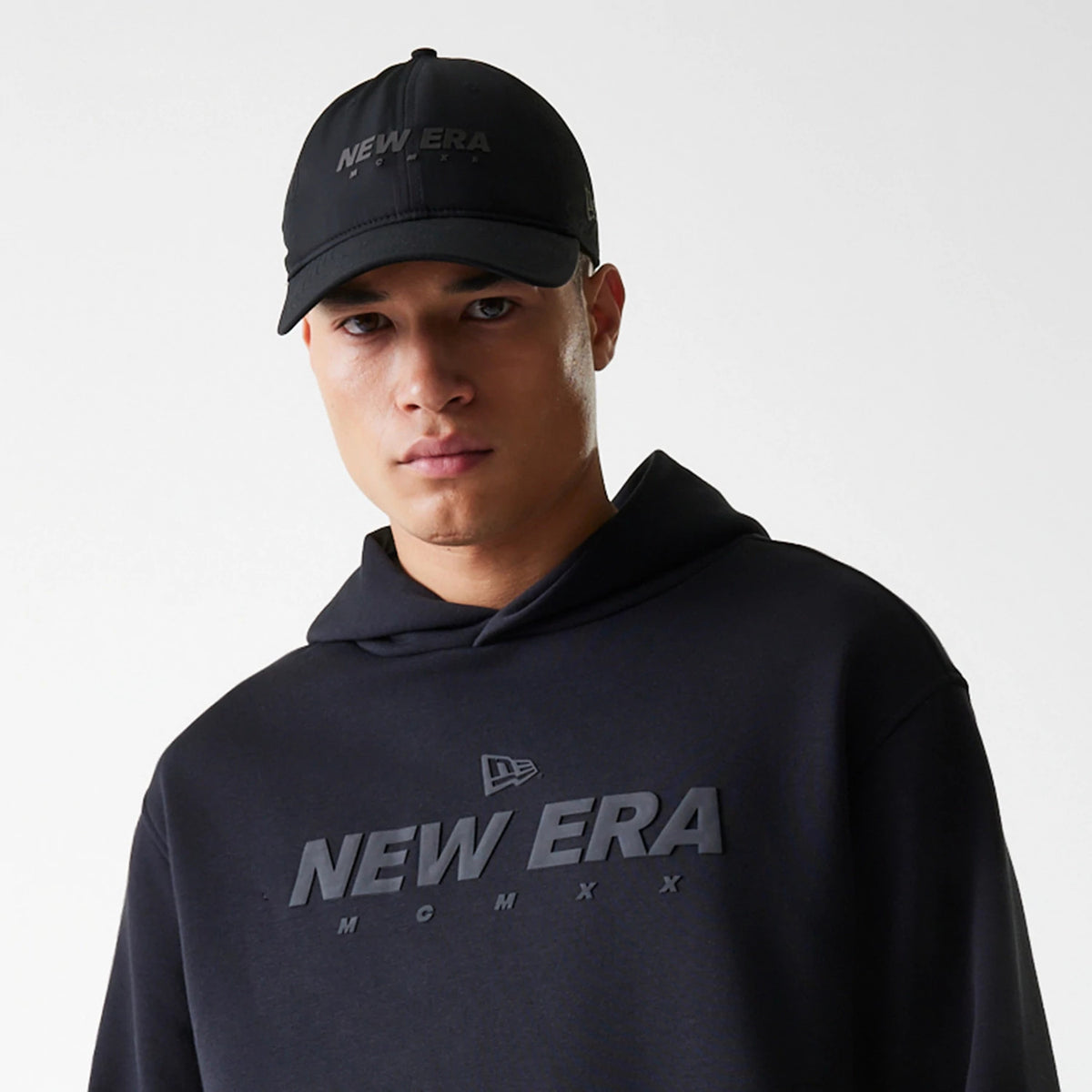 New Era Tech Black on Black Oversized Pullover Hoodie, on-model view featuring a debossed New Era logo prominently displayed on the chest and a matching black cap with a debossed New Era logo on the front above the peak.