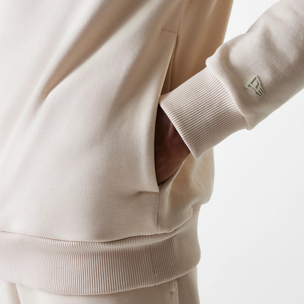 New Era Tech Light Beige Oversized Pullover Hoodie, side view highlighting the long sleeve with ribbed cuff, a hand in the side pocket, and a debossed New Era logo on the sleeve near the wrist.