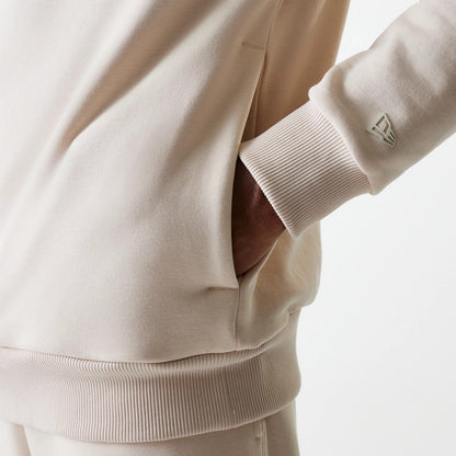 New Era Tech Light Beige Oversized Pullover Hoodie, side view highlighting the long sleeve with ribbed cuff, a hand in the side pocket, and a debossed New Era logo on the sleeve near the wrist.