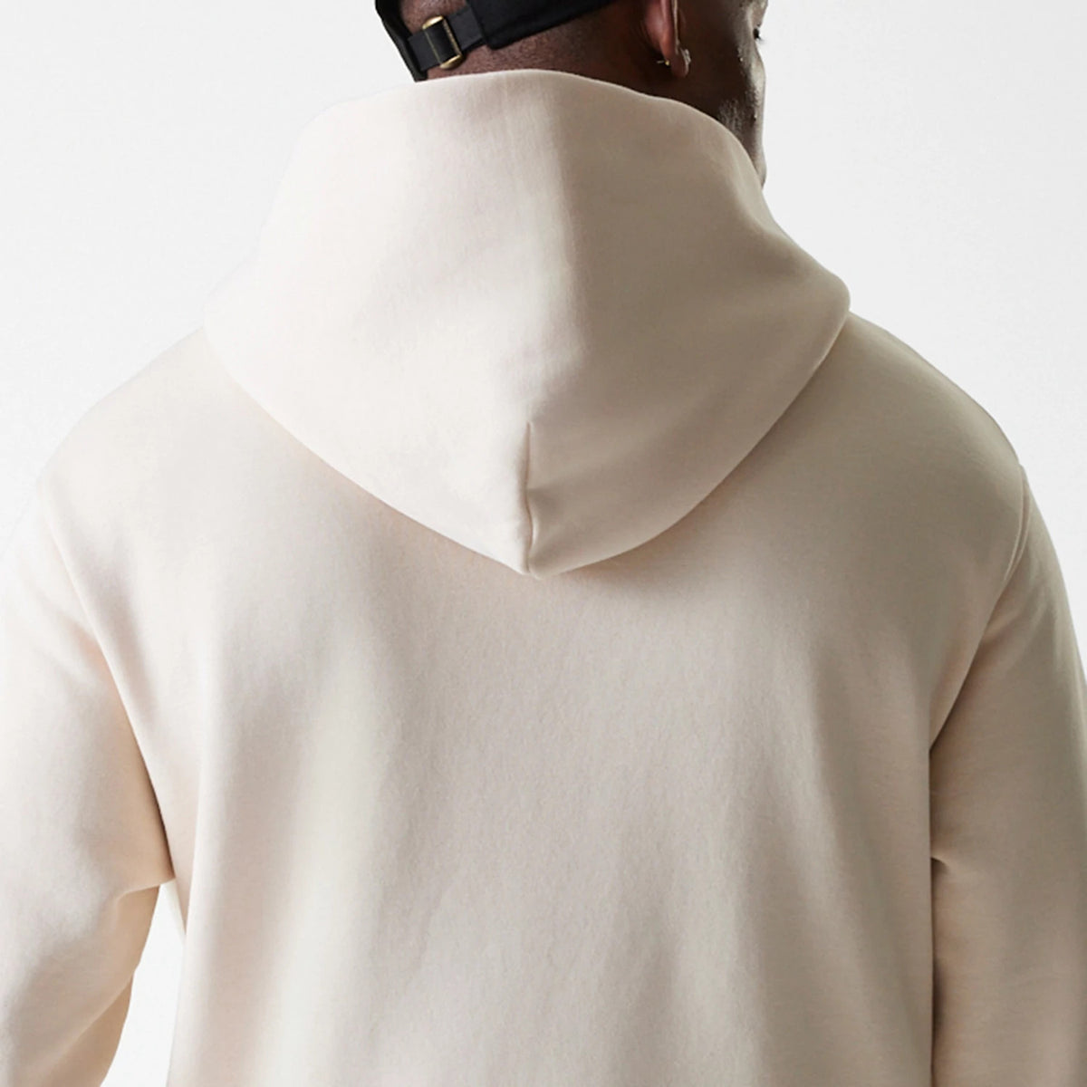 New Era Tech Light Beige Oversized Pullover Hoodie, rear view showing the hood draped down the back, a plain light beige fabric with no visible logos or designs, worn by a person with a black cap.