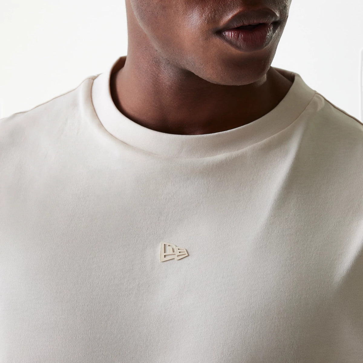 New Era Tech Light Beige Oversized T-Shirt, front view showing a raised beige New Era logo centred on the chest below the round neck, highlighting the smooth light beige fabric of the oversized design.
