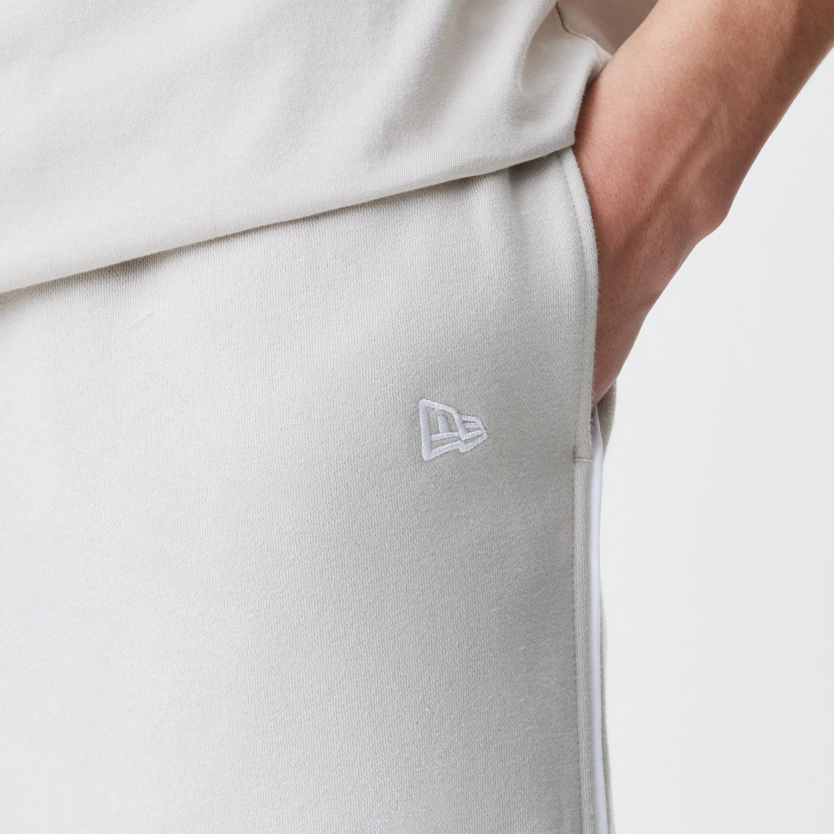 New Era LA Lakers NBA Piping Cream Joggers, close-up view showing the embroidered white New Era flag logo on the upper leg, with a white piping along the side seam and hand in pocket.