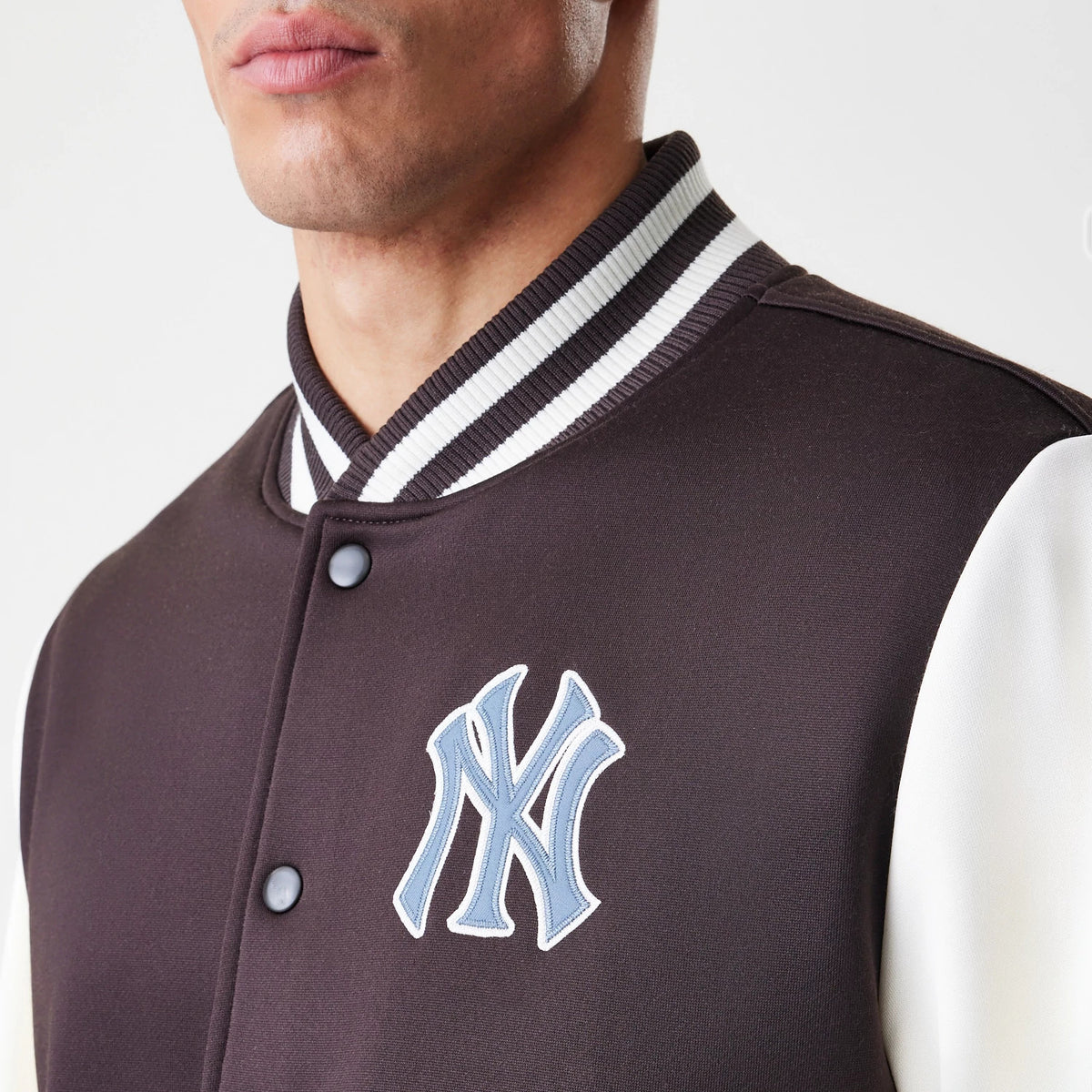 New Era New York Yankees MLB Dark Brown Varsity Jacket, front view showing a blue embroidered logo on the left chest, contrasting white sleeves, and a ribbed white and black collar, model partially wearing the jacket.