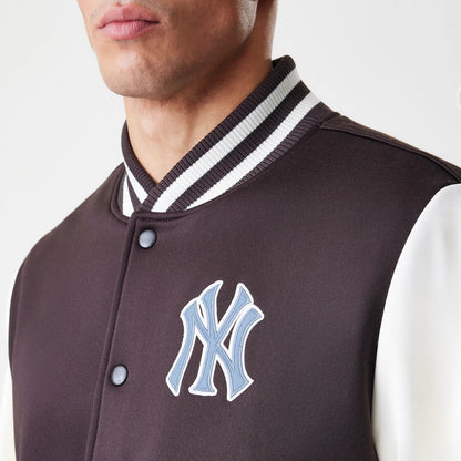 New Era New York Yankees MLB Dark Brown Varsity Jacket, front view showing a blue embroidered logo on the left chest, contrasting white sleeves, and a ribbed white and black collar, model partially wearing the jacket.