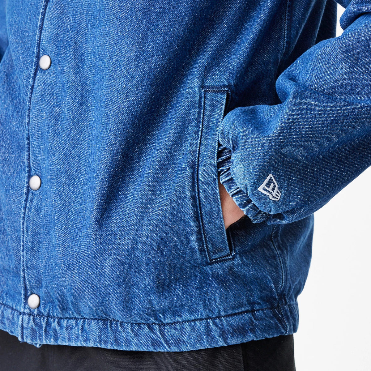 New Era New York Mets MLB Denim Dark Blue Coaches Jacket, side view showing the textured dark blue fabric, snap button fastenings down the front, a hand in the side pocket, and a white embroidered New Era logo near the cuff.