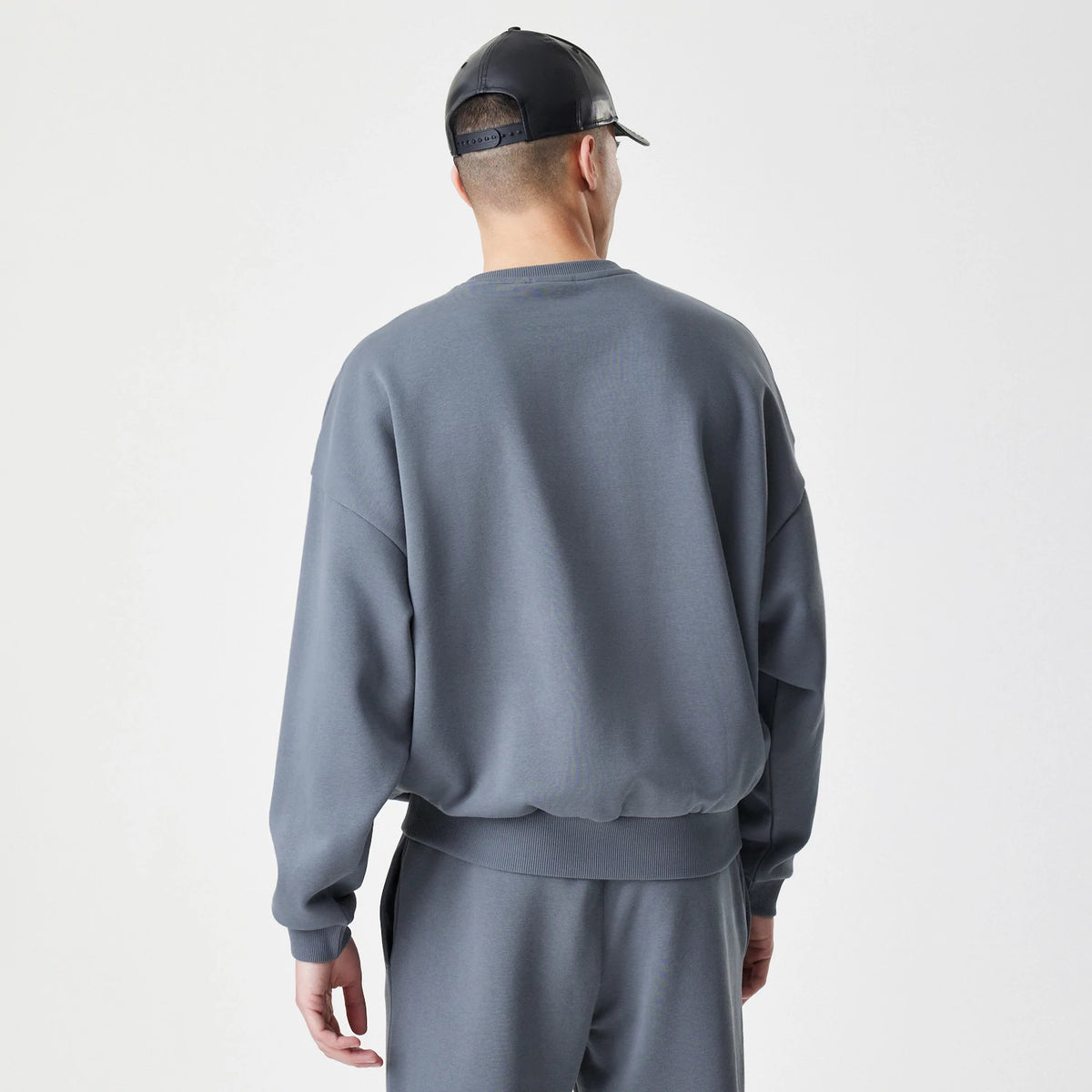 New Era LA Lakers NBA Linear Dark Grey Crew Neck Sweater, back view on a model wearing matching trousers, showing the plain dark grey fabric with ribbed waistband and cuffs, and a relaxed, loose fit.