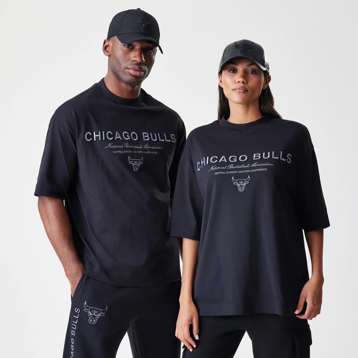New Era Chicago Bulls NBA Linear Black T-Shirt, front view on a model styled with a matching black cap, featuring the Chicago Bulls lettering and embroidered team logo on the chest, set against a plain black background.