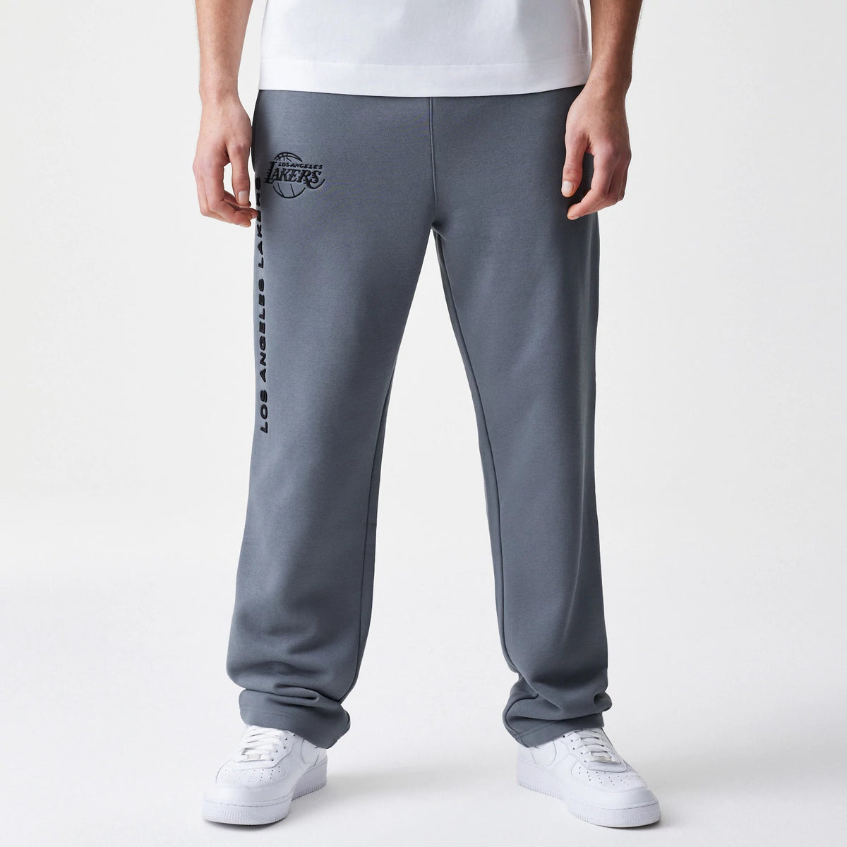 New Era LA Lakers NBA Linear Dark Grey Straight Leg Joggers, front view on a model showing the black LA Lakers logo on the left hip and Los Angeles Lakers lettering running vertically down the right leg, paired with white trainers.
