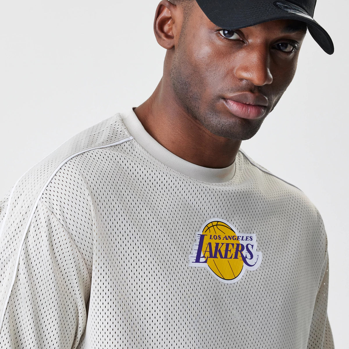 New Era LA Lakers NBA Piping Light Beige Oversized Mesh T-Shirt, front view showing the embroidered LA Lakers logo on the chest with purple lettering and a yellow basketball design, worn by a model with a black cap.
