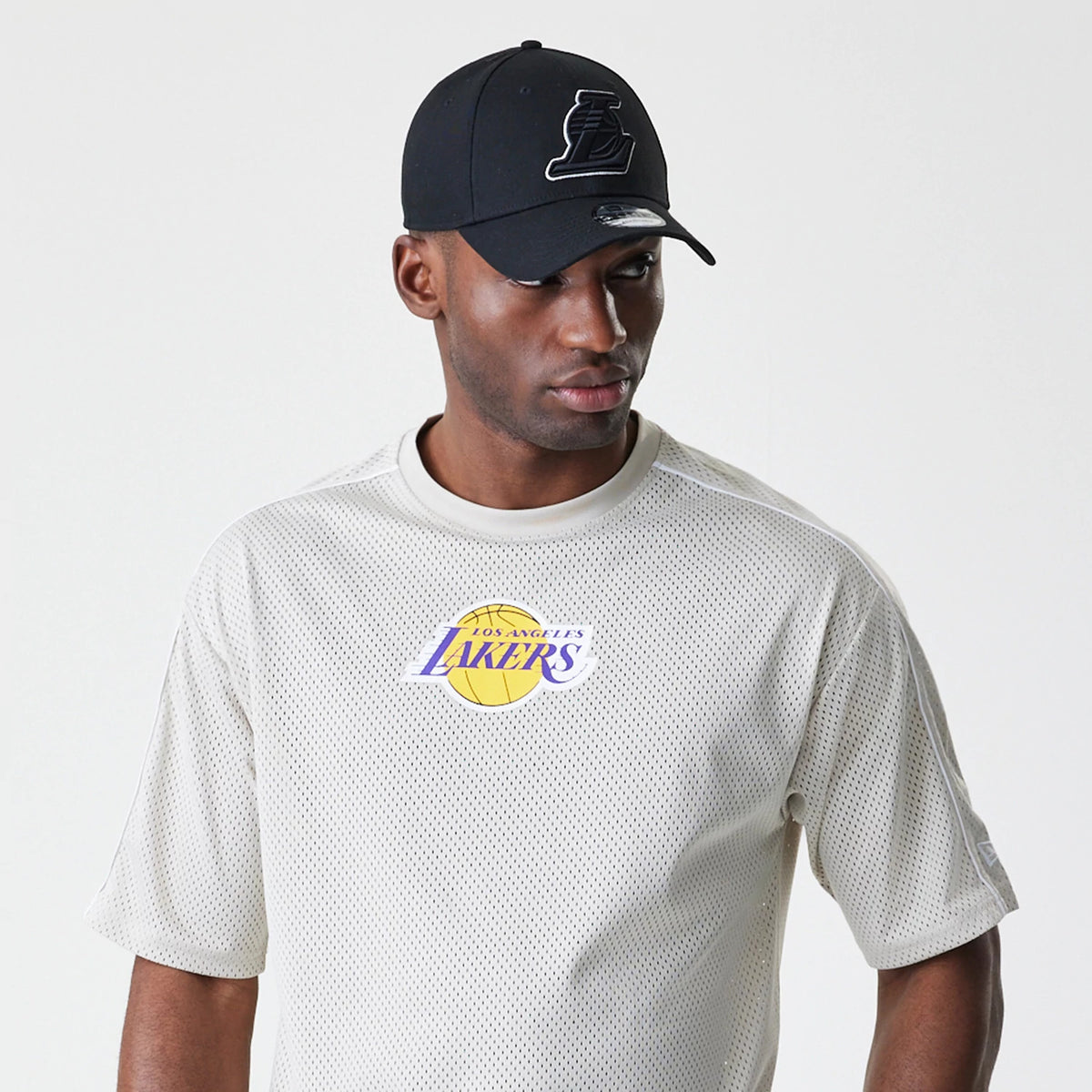 New Era LA Lakers NBA Piping Light Beige Oversized Mesh T-Shirt, front view on-model showing the purple and gold Lakers logo centred on the chest, short sleeves with a relaxed fit, paired with a black cap.