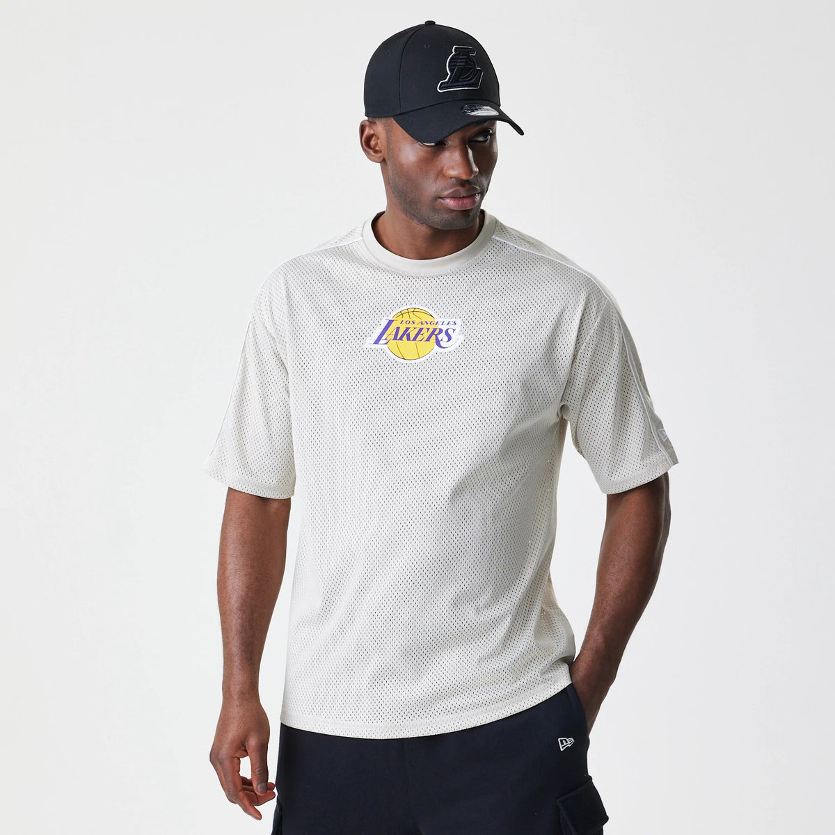 New Era LA Lakers NBA Piping Light Beige Oversized Mesh T-Shirt, front view on-model showing the purple and yellow Lakers logo centred on the chest and the shirt's loose fit, styled with a black cap featuring an embossed logo.