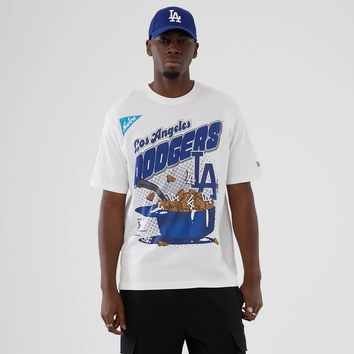 New Era LA Dodgers MLB Lifestyle Graphic White T-Shirt, front view on-model showing a Dodgers graphic featuring cereal and a spoon, New Era script on the upper left, matching blue LA cap, and blue logo printed on the sleeve.