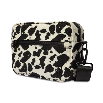 New Era Cow Jacquard Light Beige Waist Bag, side view displaying a black and white cow print pattern, a front zip pocket, a black strap with a clip, and a debossed New Era logo near the base.