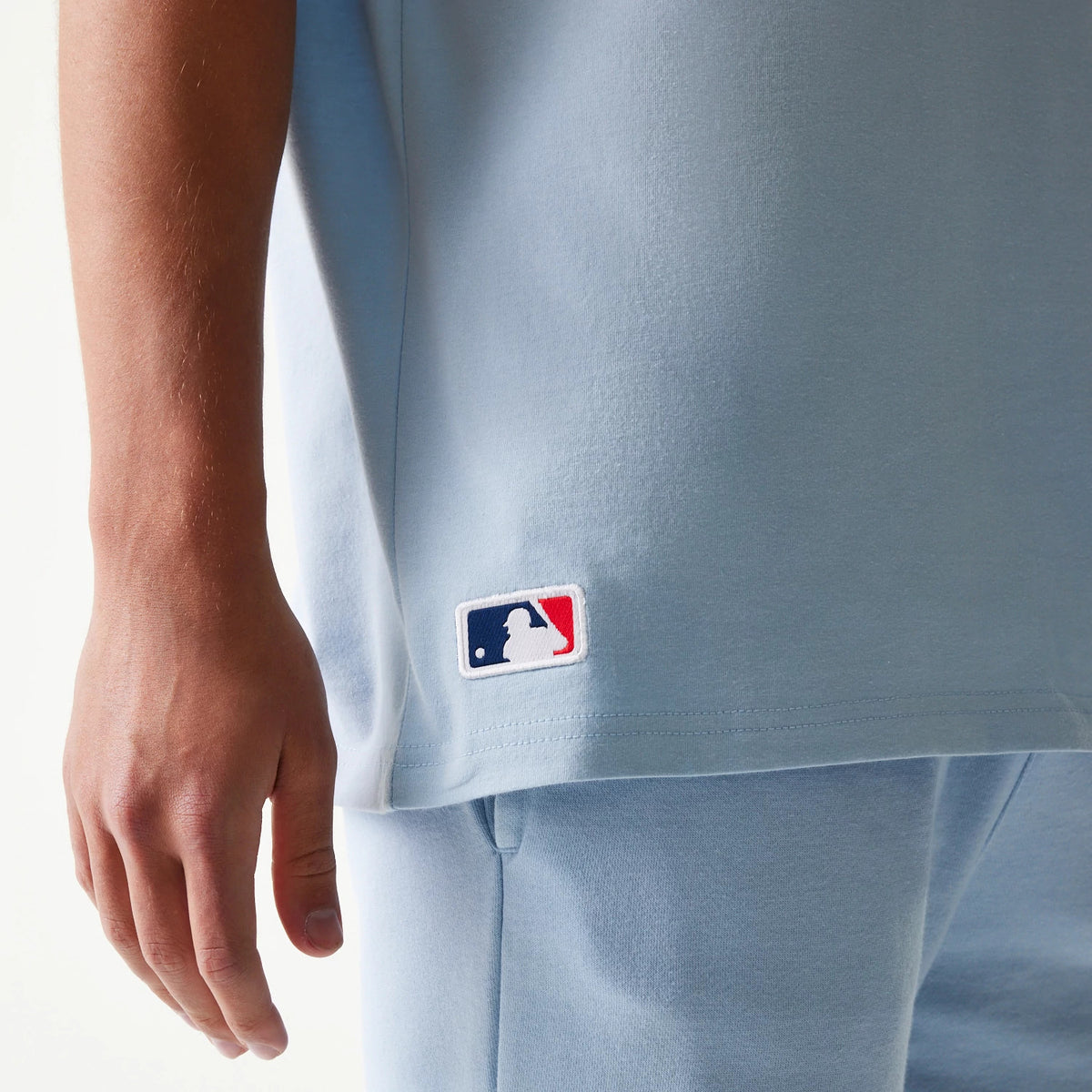 New Era New York Yankees MLB Midi Pastel Blue Oversized T-Shirt, side view showing a white embroidered MLB logo near the lower hem on the pastel blue fabric, styled with coordinated blue shorts.
