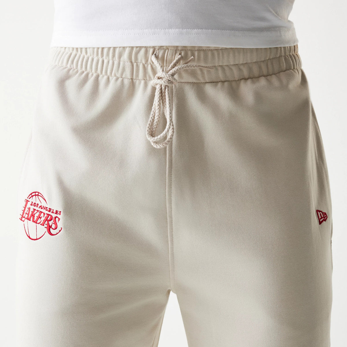 New Era LA Lakers NBA League Essential Cream Joggers, front view showing the red Lakers logo embroidery on the left leg, elasticated waistband with drawstring, and New Era flag logo on the right side.