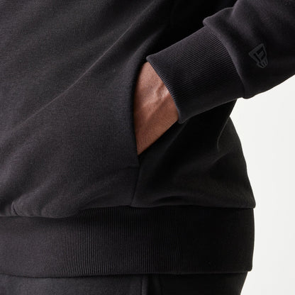 New Era Chicago Bulls NBA Black Oversized Pullover Hoodie, close-up side view showing a hand tucked into the front pocket and an embroidered New Era logo on the ribbed cuff of the black sleeve.